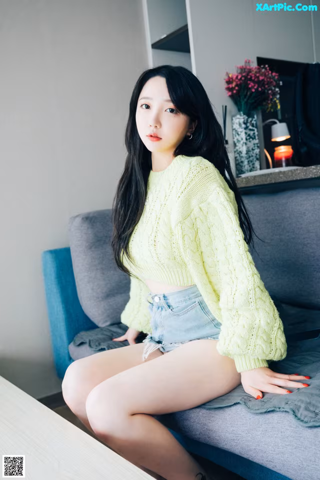 A woman sitting on a couch wearing a yellow sweater and denim shorts.